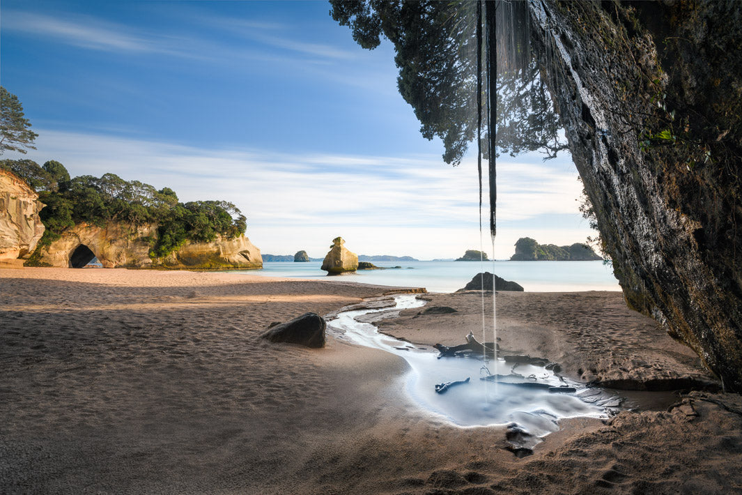 Stream of water from rocks above running into the sea at Cathedral Cove, with sand, sea and the cove in the background.