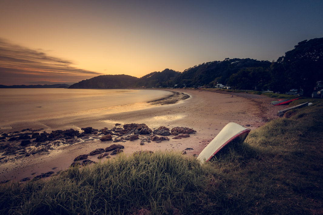 Sunrise behind hills at Long Beach in Russell, with boats leaning against the grassy dunes in the foreground, framed by trees.