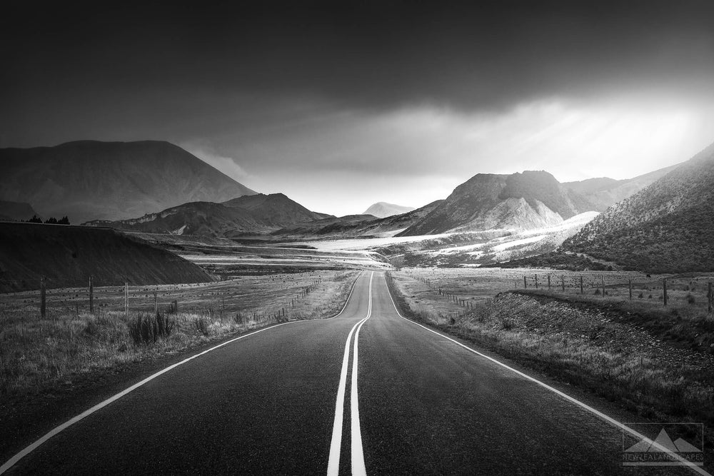 in black and white, central road leading to mountains in the background and dark stormy clouds above