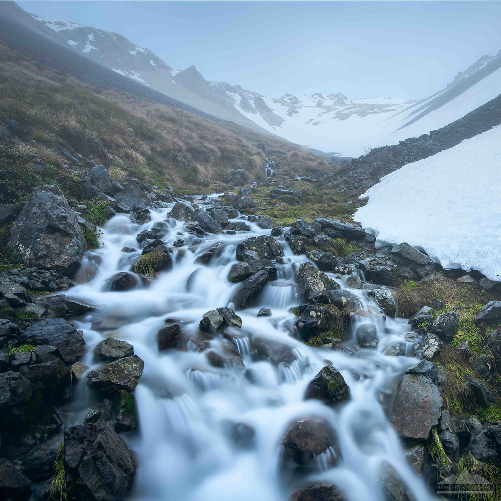 landscape photo in the snowy mountains with rocks and a stream of water tumbling in the centre and front of the photo
