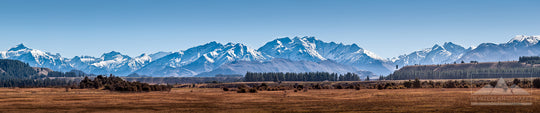 Wide panoramic photograph of New Zealand's Southern Alps with fields and trees in the foreground and clear blue sky above.