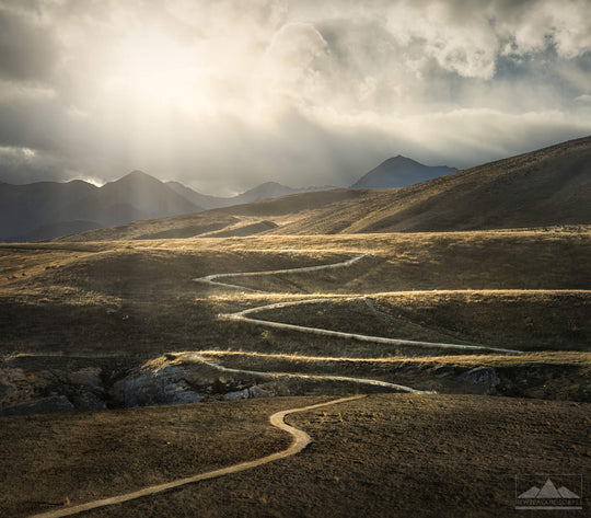 Landscape photograph of a path winding across the hillside with sunrays through the clouds above.