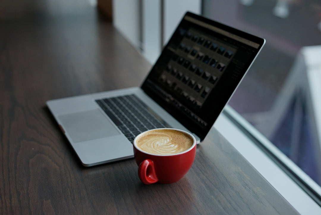Image of a coffee cup filled with a coffee and a laptop beside it, on a desk.