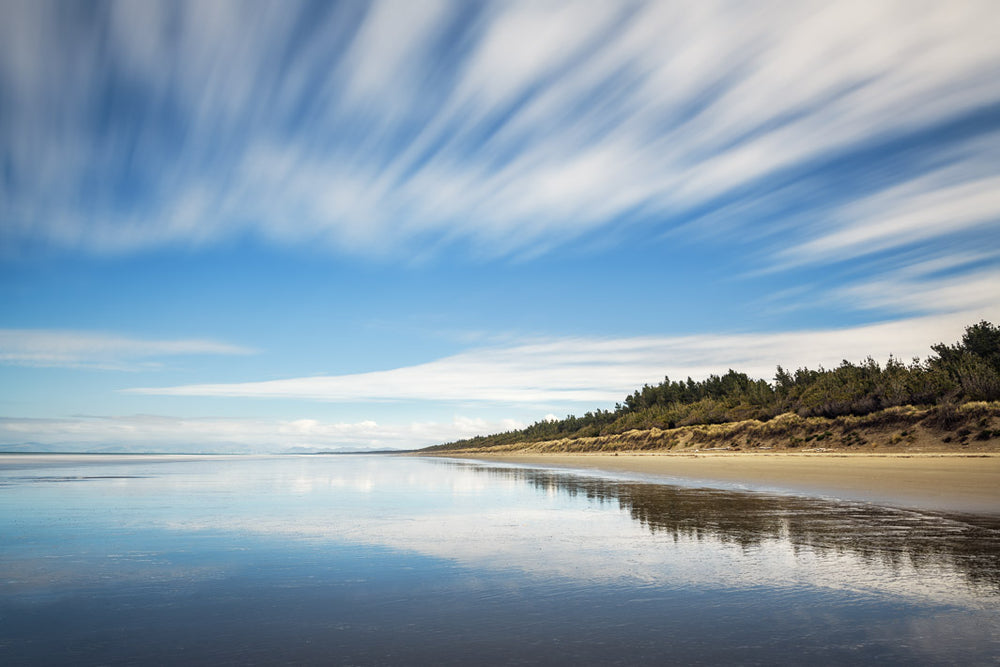Long exposure photo with clouds and trees reflected in the sea and sand below 