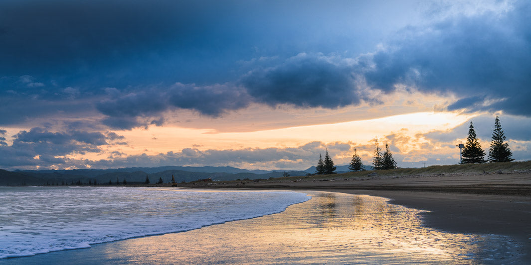 Sunset beach photo with trees and hills in the background. The sun is reflected in the shallow water below.