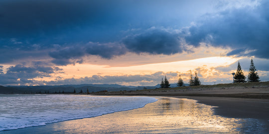 Sunset beach photo with trees and hills in the background. The sun is reflected in the shallow water below.