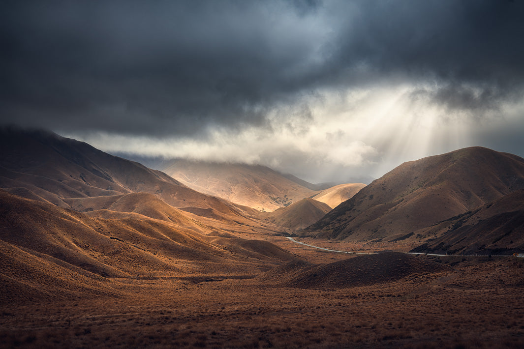 Sunrays coming through dark cloudy skies above the road and mountains of the Lindis Pass 