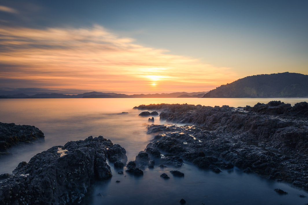 New Zealand scenic photo of the sun rising above the hills on the horizon with rocks in the foreground