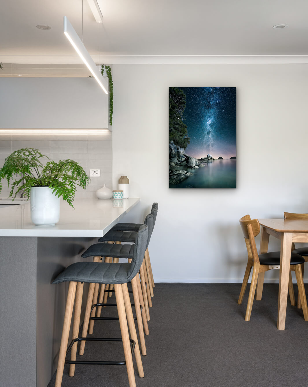 New Zealand landscape photo on canvas in modern kitchen with white bench, barstools, fern and dining setting.