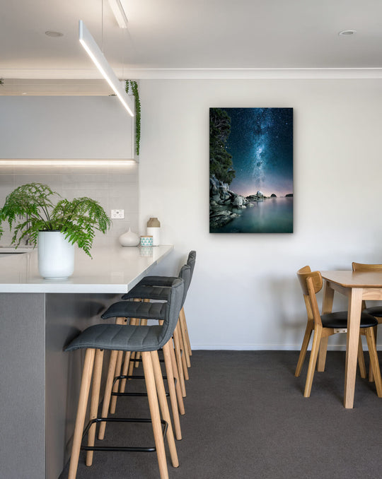 New Zealand landscape photo on canvas in modern kitchen with white bench, barstools, fern and dining setting.