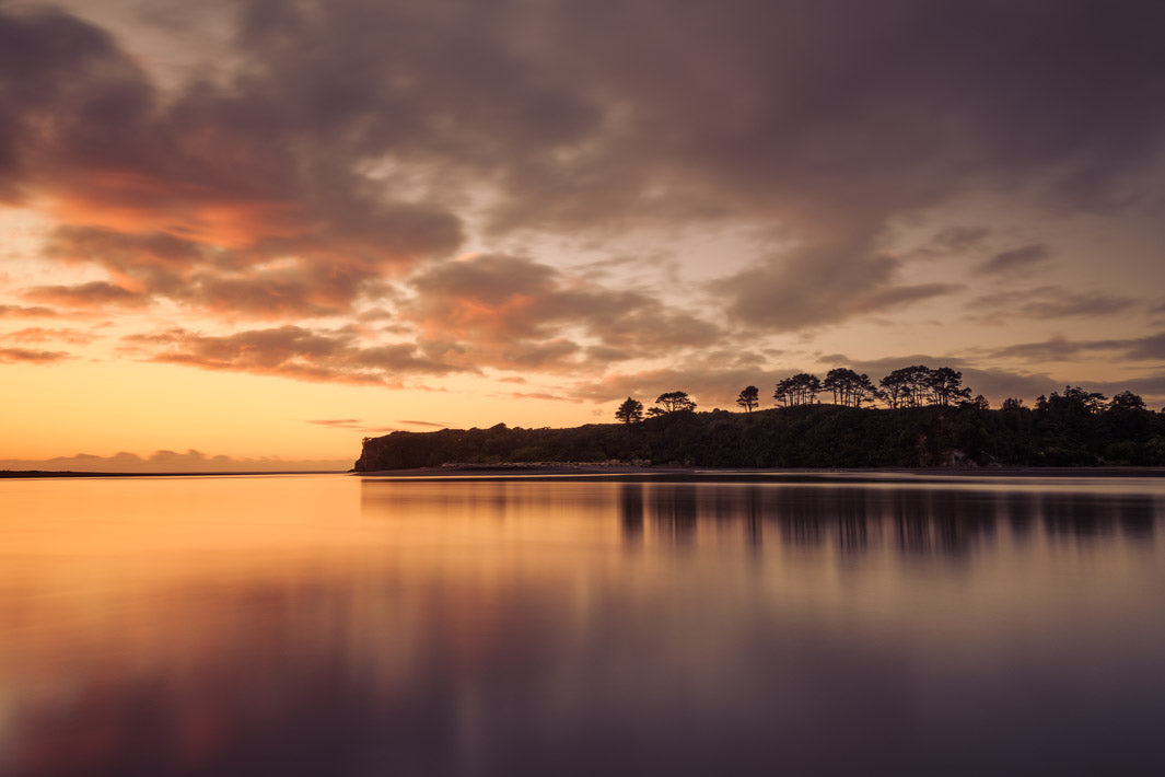 Landscape image of river mouth at sunset with trees on land in the distance. The land is reflected in the river.
