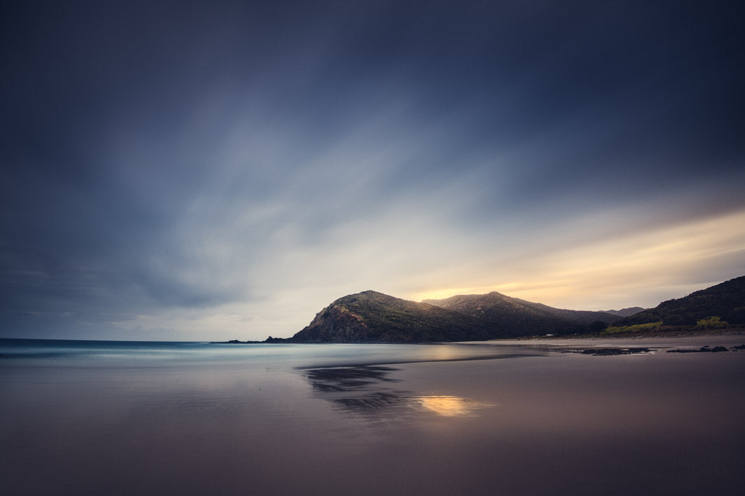 Stormy skies above a beach with the sun rising behind the hills.