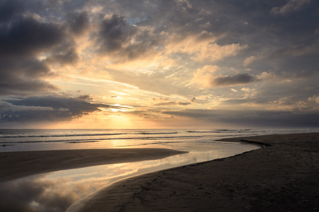 Golden sun rising through clouds and over the sea at Waihi, reflected in a stream on the sand.