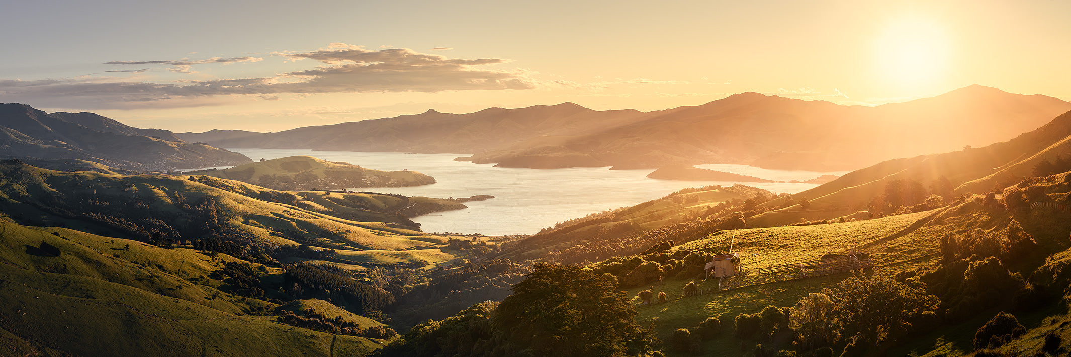 Golden sunset light over rolling green hills above Akaroa Harbour, looking toward Akaroa township and Duvauchelle from near Summit Road on Banks Peninsula.