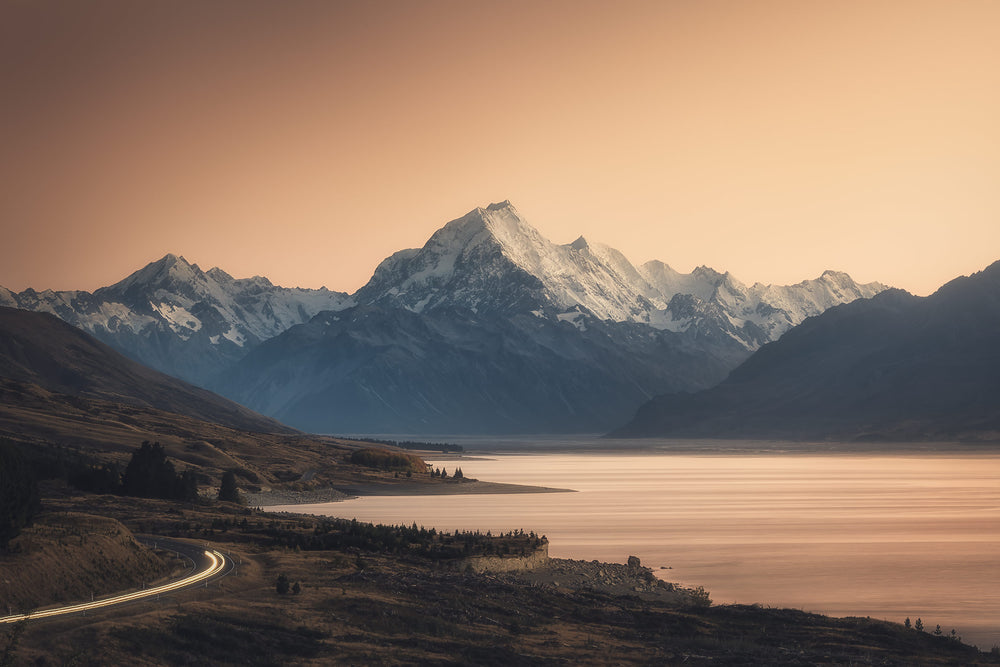 Landscape photo of Aoraki Mount Cook mountains, a lake, a car headlights on the winding road, with orange dawn colours