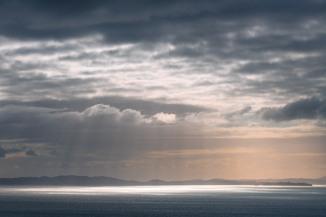 Dark cloudy skies lit by the sun beaming through over Waiheke Island and the still sea.