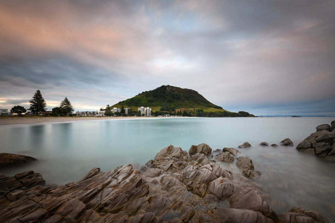 Long exposure landscape photograph of Mount Maunganui in the early morning. In the foreground are rocks from Moturiki Island.