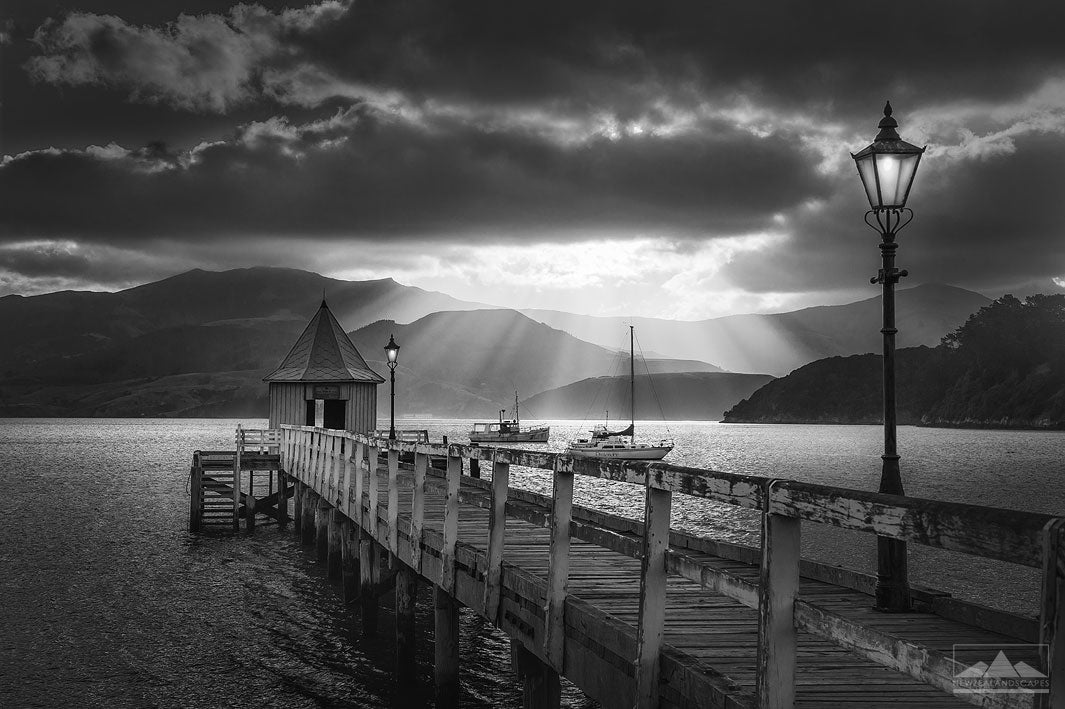 Photo in black and white of Akaroa jetty, lamp and small house at sunset with sunrays beaming down over the hills