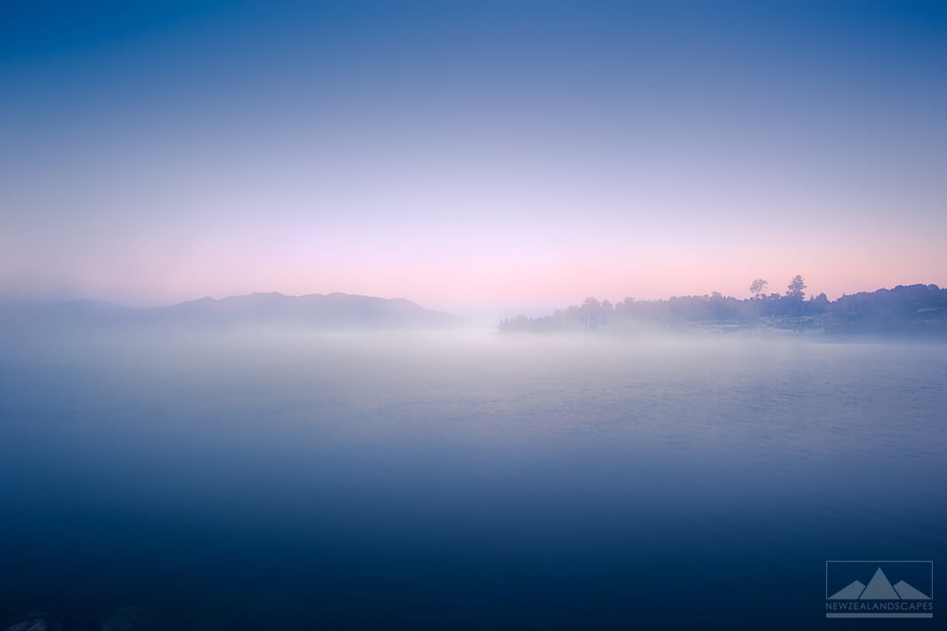 Photo of the lake at Brunner with pinks and blues in the sky with fog and mist over the blue lake.