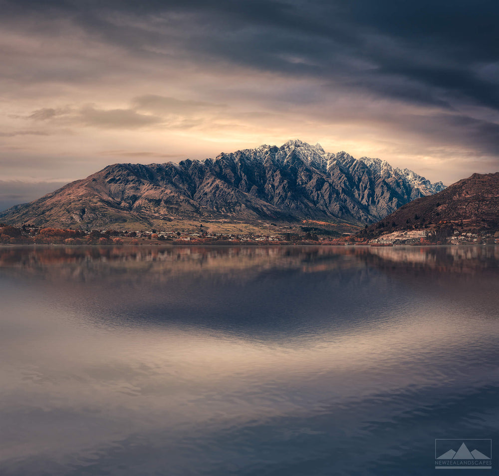 Landscape photo of The Remarkables snow capped mountain range near Queenstown with the lake in the foreground and sun rising behind clouds in the background