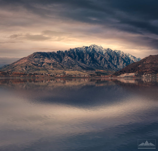Landscape photo of The Remarkables snow capped mountain range near Queenstown with the lake in the foreground and sun rising behind clouds in the background