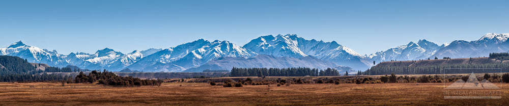 Wide panoramic photograph of New Zealand's Southern Alps with fields and trees in the foreground and clear blue sky above.