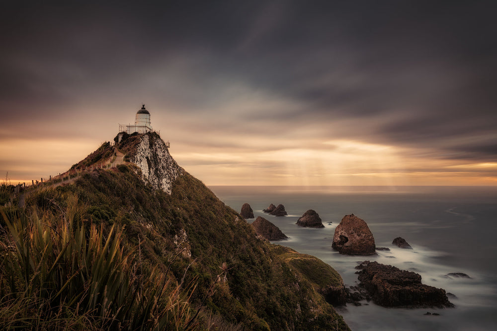 Moody skies with sunbeams through the clouds, with a lighthouse in the foreground, at the top of a rugged grassy cliff. The sea and rocks are below.