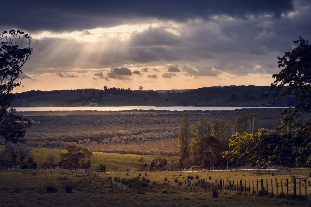 Sunbeams through the clouds over Kaipara Harbour, overlooking fields of grass and trees.