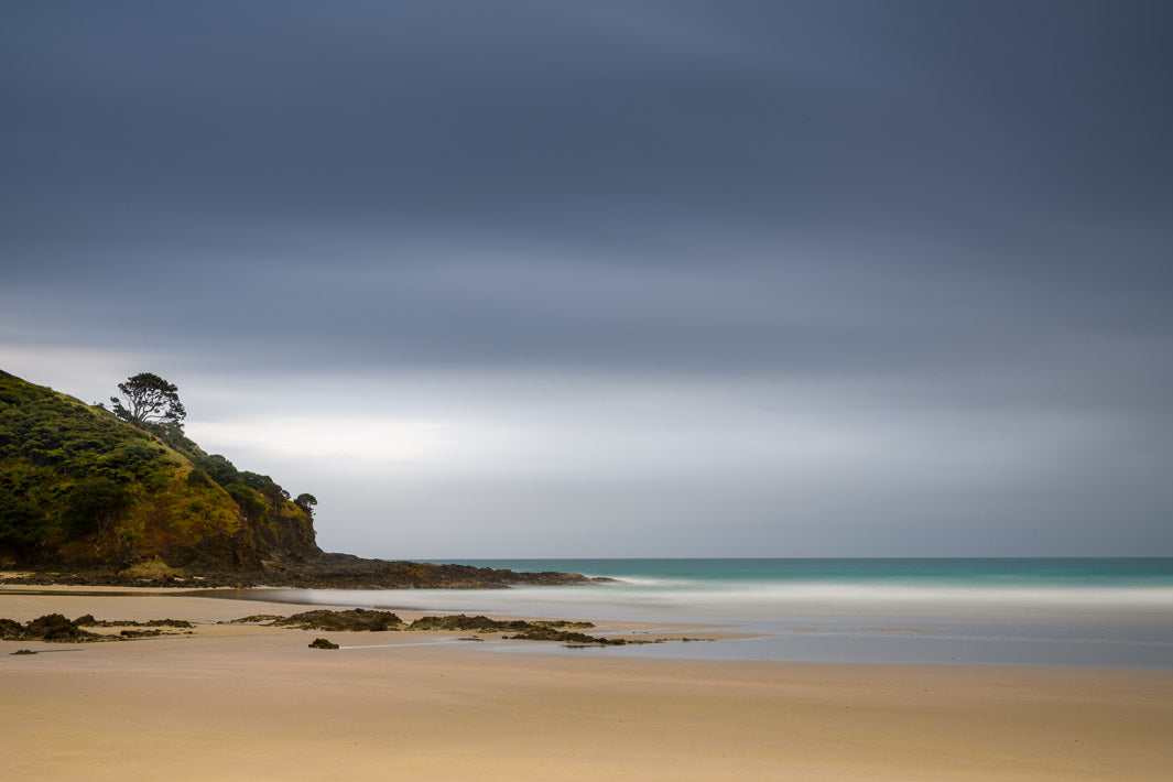 Golden sands beach with stormy grey skies above, tree on the hillside and smooth long exposure sea.