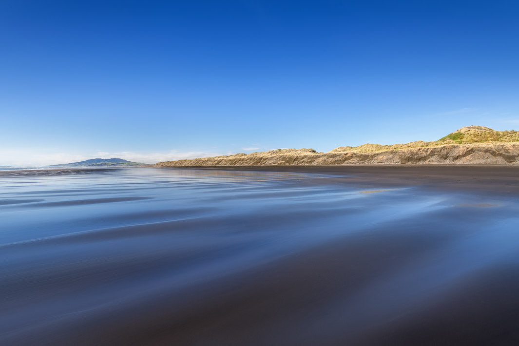 Smooth waves lead the eye to sand dunes with a clear blue cloudless sky above.