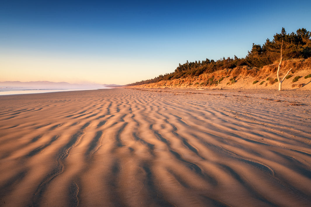 Large stretch of rippled sand with dunes and clear blue skies above.