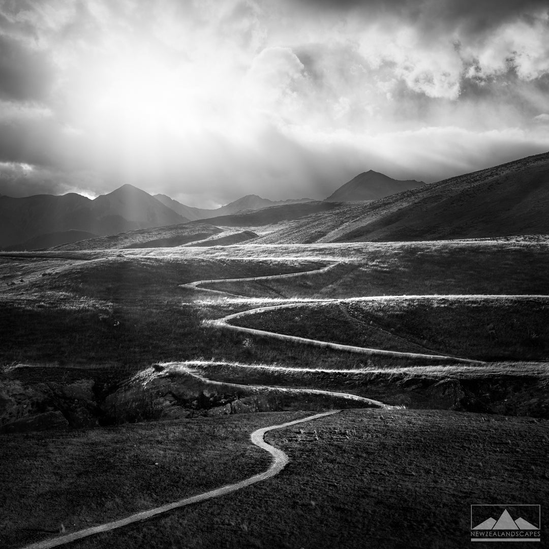 Farming path or track meandering through the New Zealand rolling hills with mountains and sunrays in the background