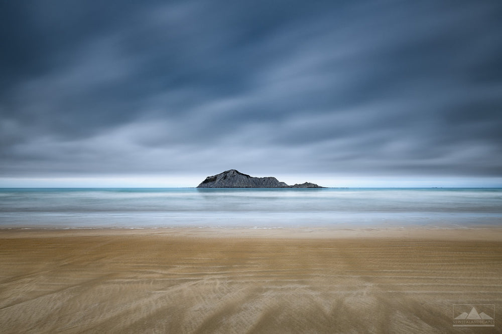 Long exposure photo of Bare Island off the coast of Waimarama Beach, with moody dark clouds in the sky and golden sand in the foreground