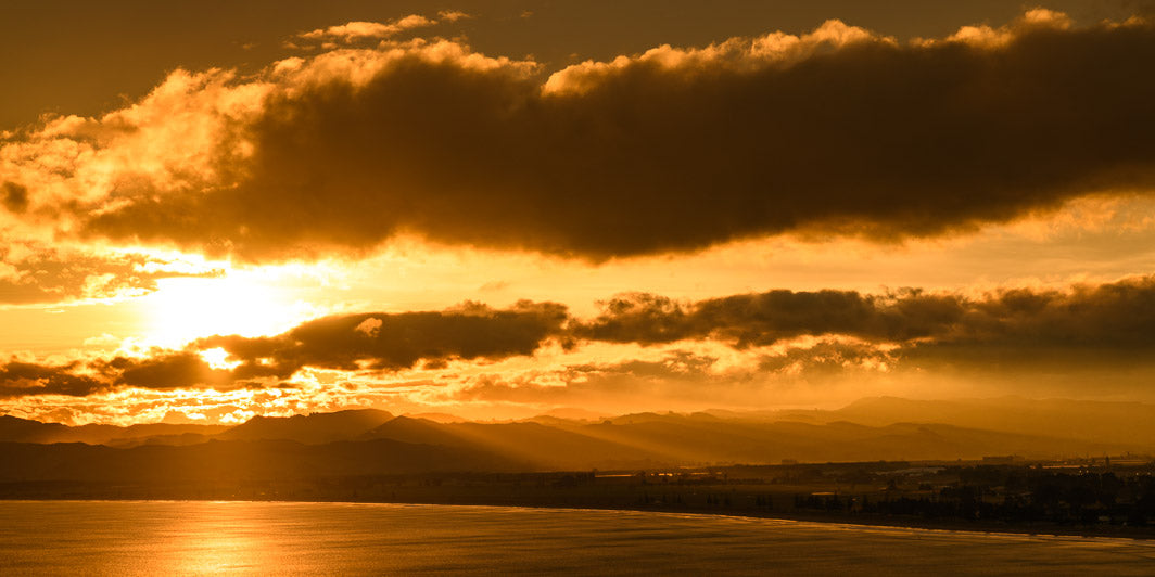 Panoramic landscape photo of a golden yellow sunset in Gisborne. Photo depicts setting sun through clouds with mountains, land and sea below in the distance.