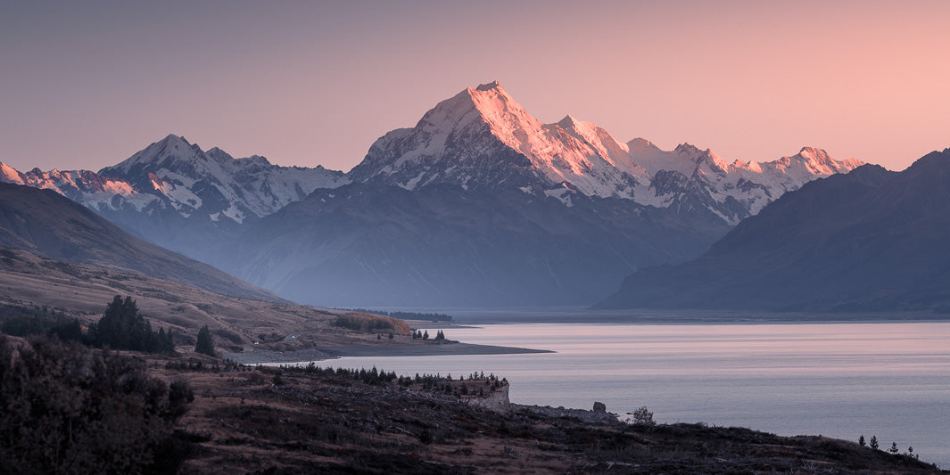 Panoramic photo of New Zealand's highest mountain Aoraki Mount Cook with the sun highlighting the snowy peaks with a lake in the foreground