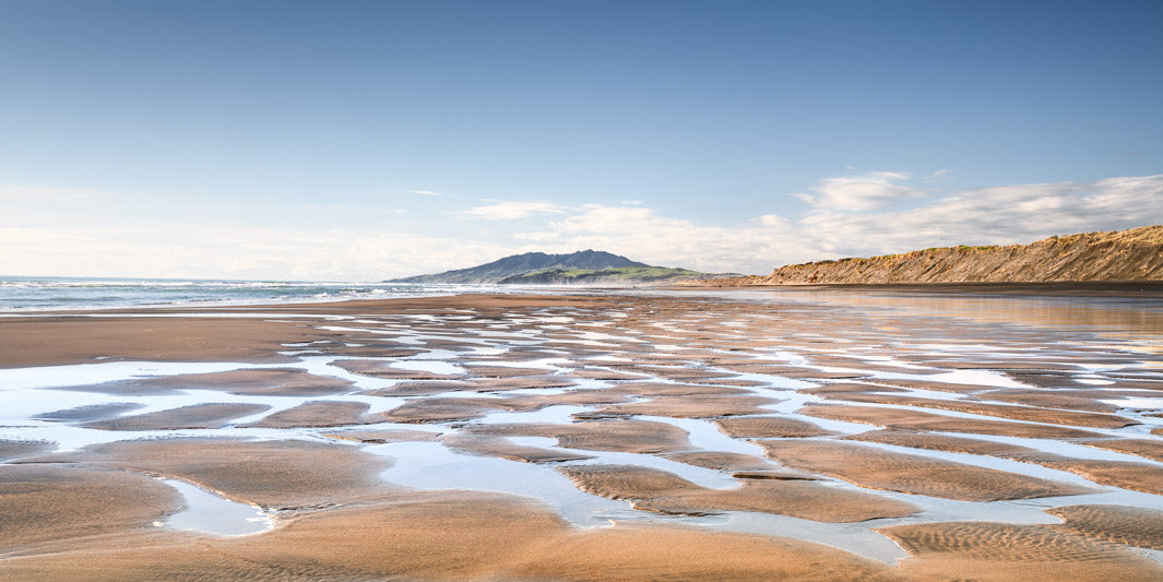 Panoramic photo of rippling sand and pools of hot sea water at New Zealand's Kawhia, with a hill in the distance and sand dunes to the right.