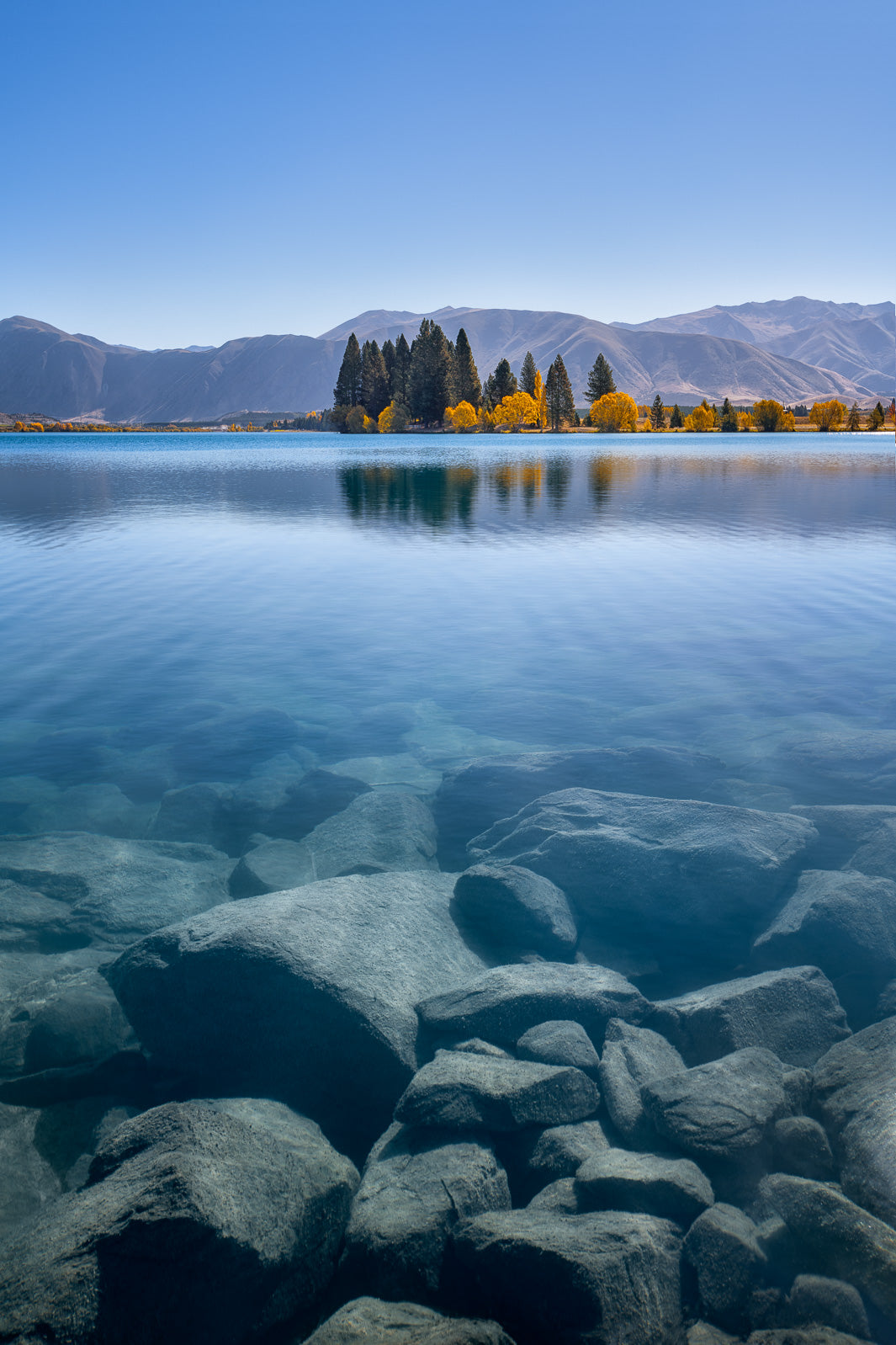 Photo of a New Zealand lake with rocks visible through the clear water in the foreground and trees and mountains in the background of a clear Canterbury blue sky.
