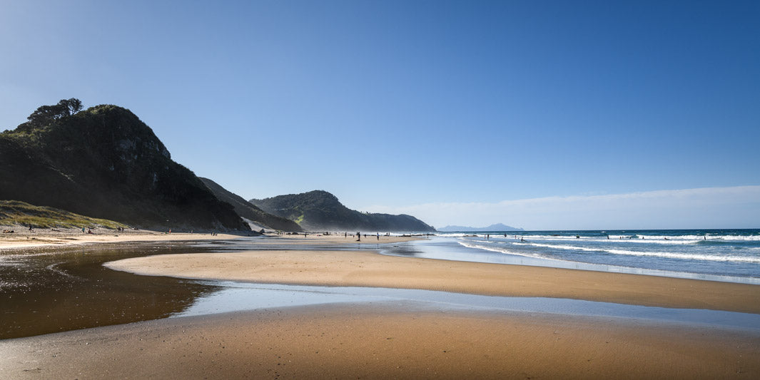 Landscape photo of a beach in Northland, New Zealand, with the sea waves to the right and hills to the left and clear blue sky.