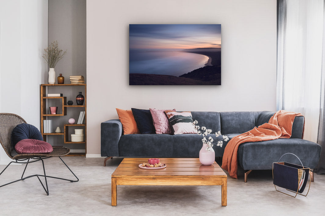 Sweeping sea view photograph hung on a wall above a couch, coffee table and chair, in front of a bookcase.