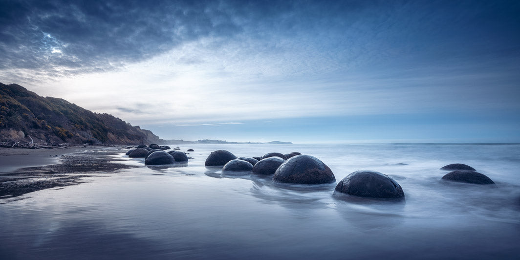 Large round boulders in the sea, with a long exposure capture, with hills in the background and soft clouds in the sky.