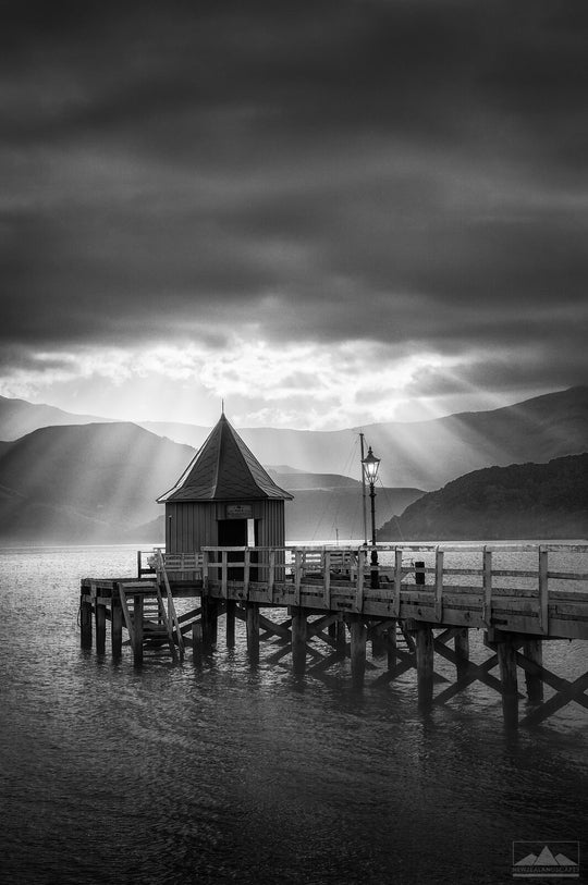 Jetty in Akaroa Harbour with sun rays coming out from a cloudy sky.