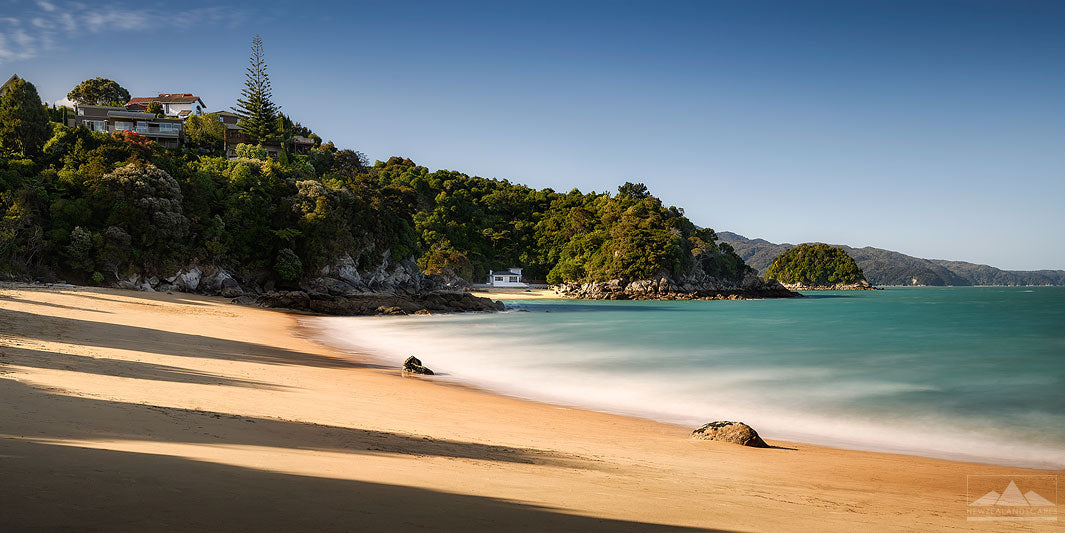 Long exposure landscape photo of Breaker Bay with golden sand and blue sea highlighted by the sun with trees and houses on a hill in the background. 