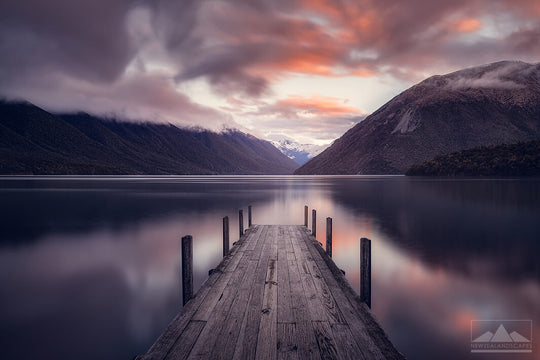 NZ landscape photo of Lake Rototi at sunset, with the jetty in the foreground, mountains reflected behind and pink clouds above.