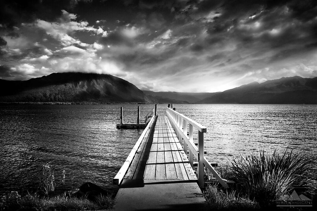 Black and white photo of a cloudy Lake Brunner with a jetty in the centre of the image and hills in the background