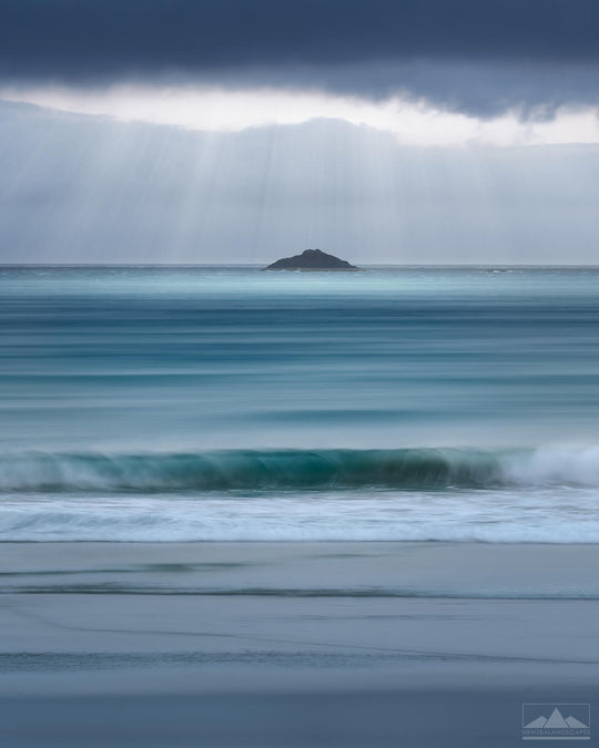 Vertical photo wall art of stormy skies, sunbeams over a rock in the sea and waves rolling in