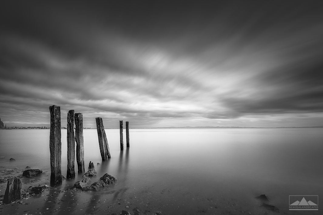 long exposure black and white image ofwooden jetty remnants in the sea with heavy clouds above