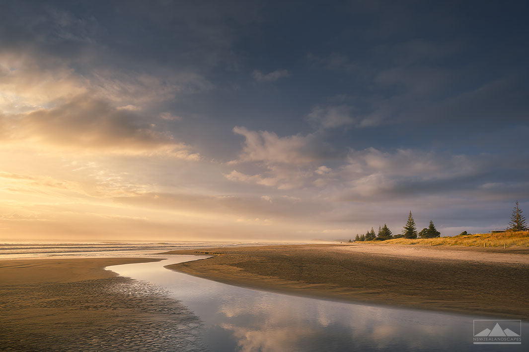 Landscape photograph of Waihi Beach at sunrise with the clouds reflected in the stream in the foreground