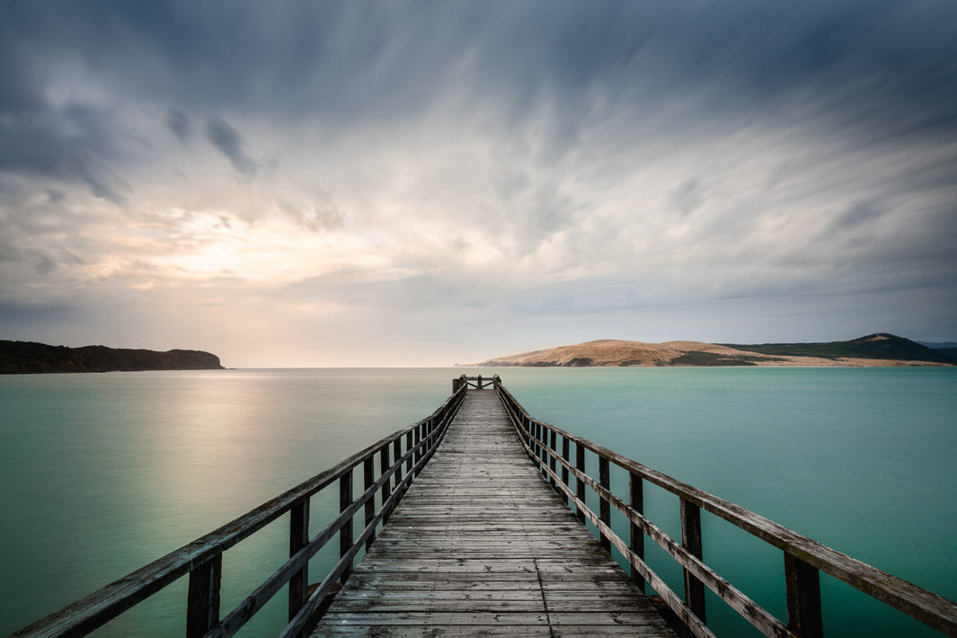 Long exposure photo print of a wooden wharf in a harbour, with hills surrounding it.