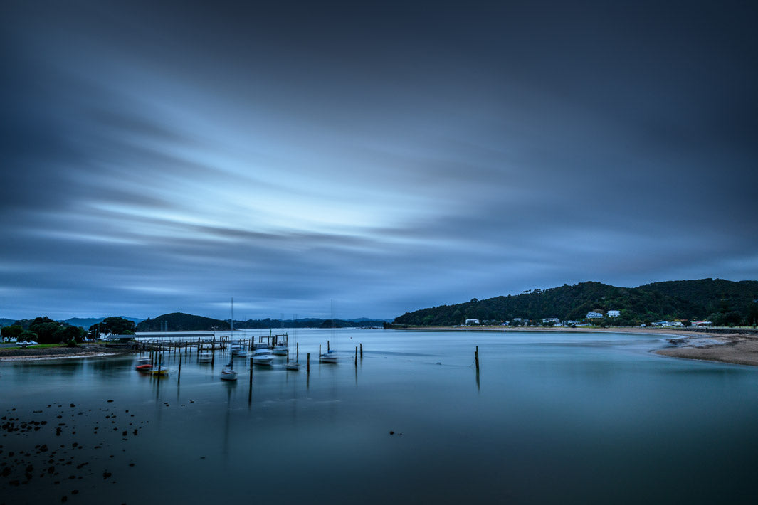 Evening long exposure capture of a bay with small boats, hills and distant houses.