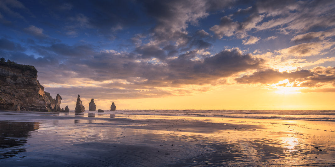 New Zealand panoramic image of the Three Sisters rock formation in the sea with the sun setting behind clouds in the sky